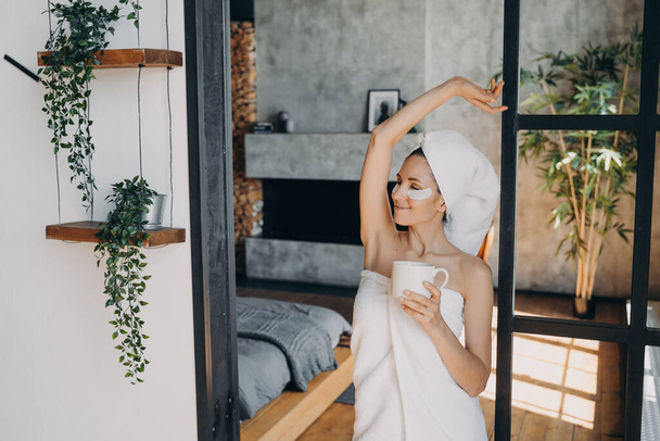 Girl applies anti age eye patches and smiling. Weekend morning at home. Young lady with cup of coffee relaxing at luxurious spa resort. Beautiful european woman wrapped in towel after bathing. - Valokuva, kuva