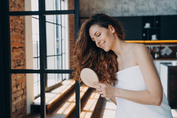 Hygiene and hair care concept. Young european woman combs her long gorgeous curly hair with brush. Happy girl wrapped in towel doing hair therapy in modern room of apartment or hotel. - Foto, Bild