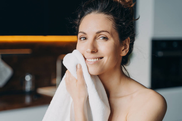 European girl is wiping face with towel after washing. Young brunette woman takes shower at home and doing skin care. Hygiene and freshness, dermatology and spa procedures. - Fotoğraf, Görsel