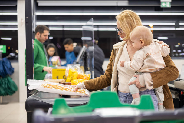 Мама ходила по магазинам со своим маленьким мальчиком, держала ребенка, пока клала продукты в кассу в продуктовом магазине - Фото, изображение