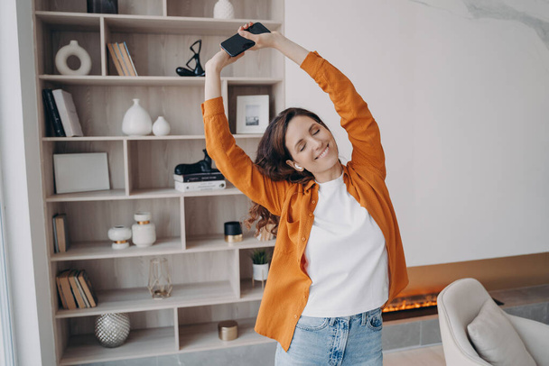 Happy european woman is dancing alone at home. Girl in earphones is having fun in her living room. Music listening online. Concept of weekend, relaxation and emotion expression. Cosy home interior. - 写真・画像