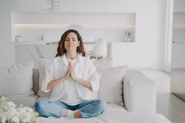 Serene latin woman at home. Posture exercise, lotus asana. Young european woman is practicing yoga and meditation on couch with her legs crossed. Minimal white modern scandinavian interior. - Valokuva, kuva