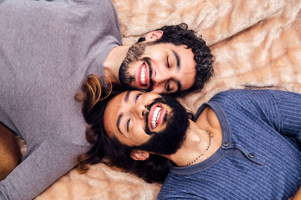 top view of a happy gay male couple laughing lying on the bed with their faces together, concept of real people lifestyle at home and love between people of the same sex - Zdjęcie, obraz