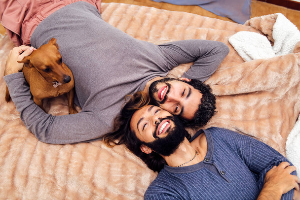 happy gay male couple smiling lying with their little dog on the bed with faces together, concept of real people lifestyle at home and love between people of the same sex - Foto, afbeelding