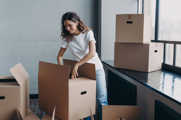 Happy young european woman unpacking boxes. Girl near window in white t-shirt indoors. Woman is opening cardboard box and smiling. Relocation and independence concept. - Fotó, kép