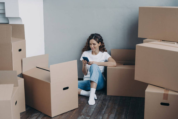 Happy young european woman with smartphone is packing boxes. Girl is chatting on mobile phone and sitting on the floor. Easy moving and shipping service order concept. - Foto, immagini