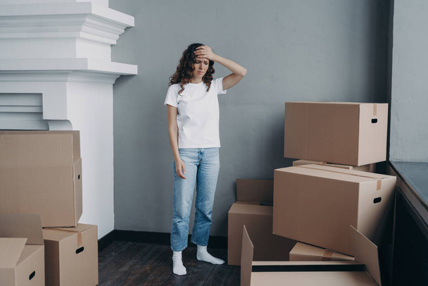 Tired girl is moving. Woman getting headache from boxes packing. Annoyed young lady in jeans and white t-shirt is going to relocate. Homeowner gathering things and getting away from luxurious house. - Foto, Imagen
