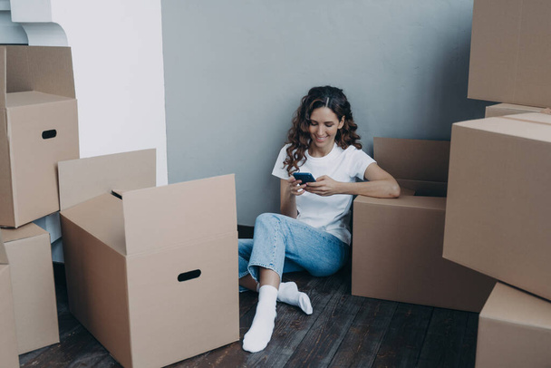 Girl is texting on mobile phone and sitting on the floor. Happy attractive spanish woman surrounded with cardboard packages and boxes. Easy moving to new apartment. Shipping service order concept. - Fotografie, Obrázek