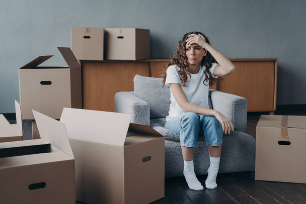 Headache from moving. Exhausted woman sitting among packed cardboard boxes. Spanish young lady in jeans and white t-shirt going to relocate. Depression and stress from moving concept. - Foto, afbeelding