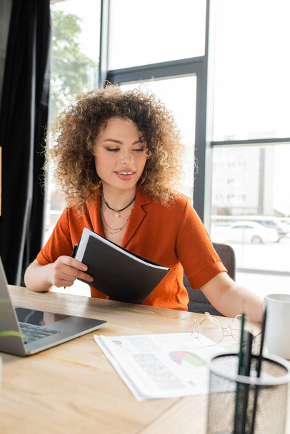 satisfied businesswoman with curly hair holding folder near laptop on office desk  - Foto, afbeelding
