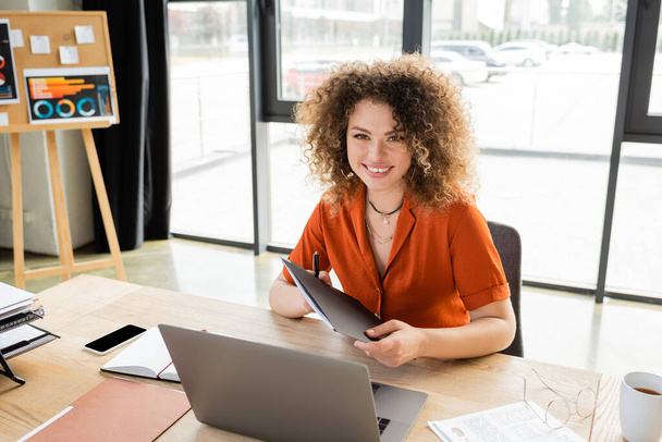 cheerful businesswoman with curly hair holding folder near laptop and infographics on office desk  - Foto, imagen