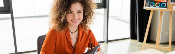 fröhliche Geschäftsfrau mit lockigem Haar hält Ordner in modernem Büro, Banner  - Foto, Bild