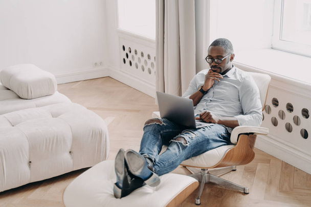 Confident professional at work in front of laptop. Relaxed african american businessman in formal wear is sitting in cosy chair at home. Online freelance job, startup project, remote work. - Foto, Imagen