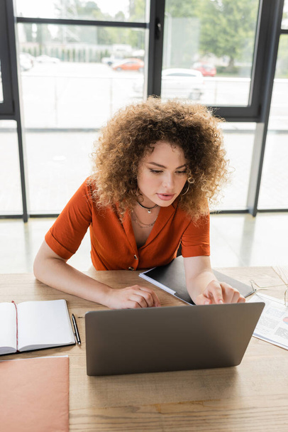 visão de alto ângulo de mulher de negócios encaracolada usando laptop perto de notebook em branco na mesa  - Foto, Imagem