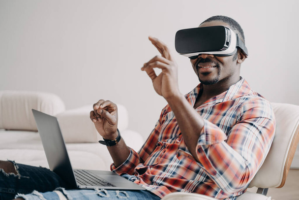 Distant work and futuristic gaming. African american man in vr goggles working with laptop at home. Relaxed freelancer clicks virtual buttons. Internet technology for business and entertainment. - 写真・画像