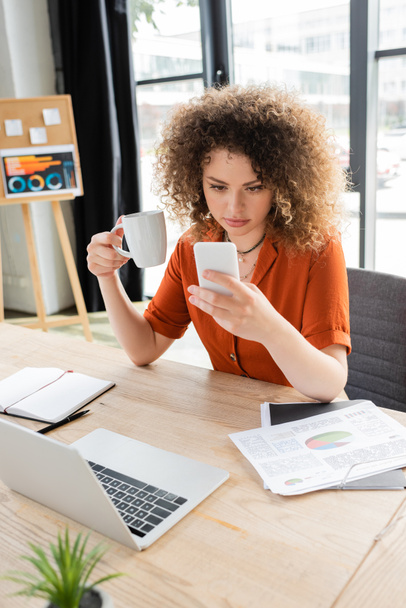 joven mujer de negocios con cabello rizado sosteniendo taza de té y mirando el teléfono inteligente cerca de la computadora portátil en el escritorio  - Foto, Imagen