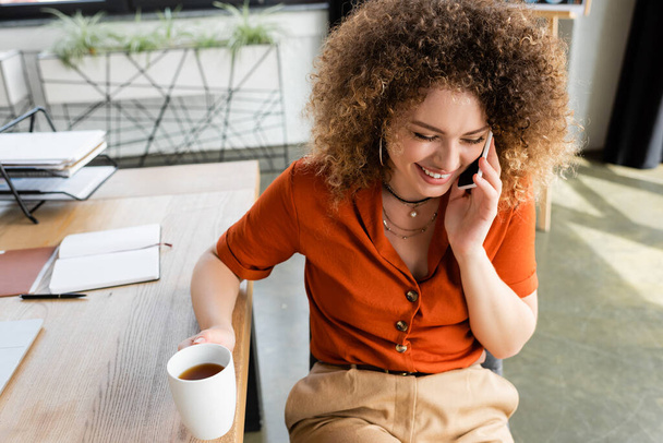 Blick von oben auf freudige Geschäftsfrau, die mit dem Smartphone spricht und eine Tasse Tee im Büro hält  - Foto, Bild