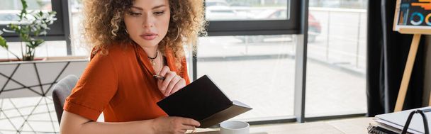 concentrated businesswoman holding notebook and pen near cup of tea in office, banner  - Фото, изображение