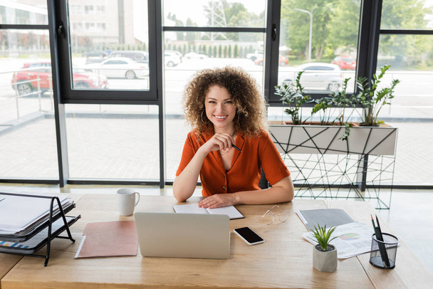 pleased businesswoman with curly hair looking at camera near gadgets and documents on desk  - Фото, зображення