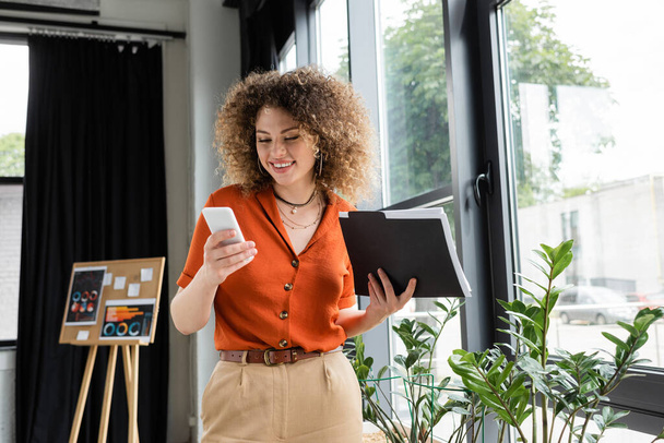 cheerful businesswoman with curly hair holding folder and using smartphone in office  - Fotoğraf, Görsel
