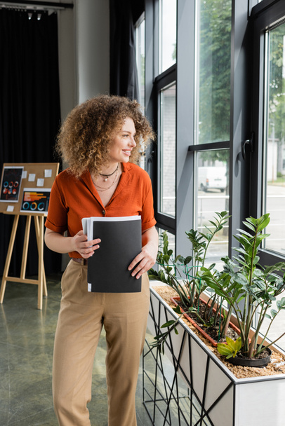 happy businesswoman with curly hair holding folder and looking at window in modern office  - Fotografie, Obrázek