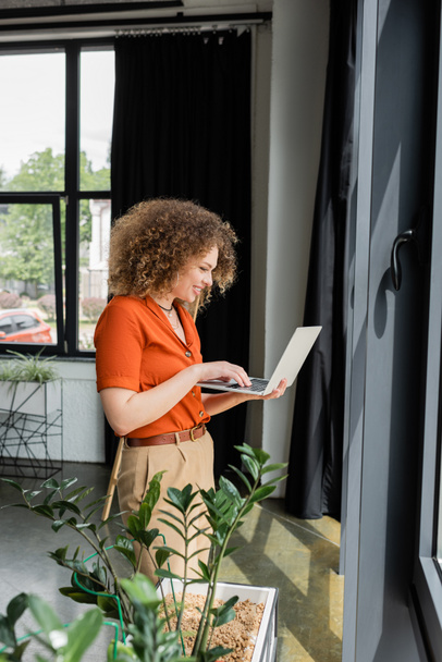 side view of happy businesswoman with curly hair using laptop next to green plants in modern office  - Fotografie, Obrázek