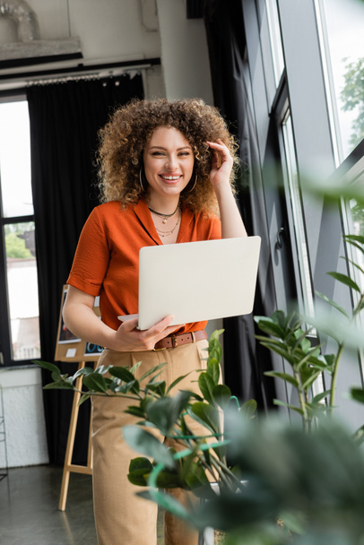 positive businesswoman with curly hair holding laptop next to green plants in modern office  - Zdjęcie, obraz