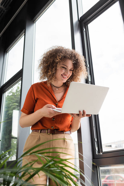low angle view of happy businesswoman with curly hair looking at laptop while standing near windows in office  - Foto, afbeelding