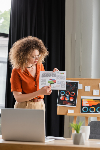 krullend zakenvrouw in bril wijzend met pen op infographics terwijl het maken van online presentatie tijdens videogesprek  - Foto, afbeelding