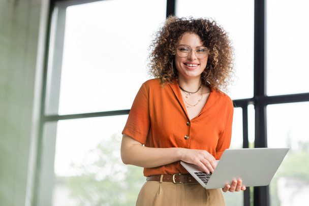 curly businesswoman in glasses smiling while holding laptop in office  - Photo, Image