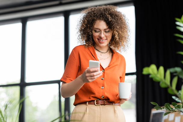 alegre mujer de negocios en gafas de mensajería en el teléfono inteligente mientras sostiene la taza de café en la oficina moderna  - Foto, Imagen