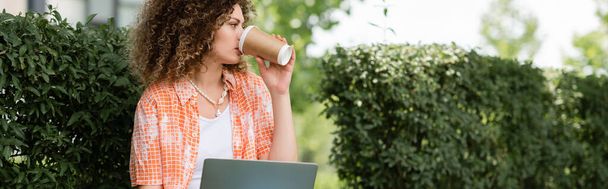 молодая женщина с вьющимися волосами пьет кофе, чтобы пойти и использовать ноутбук на открытом воздухе, баннер  - Фото, изображение