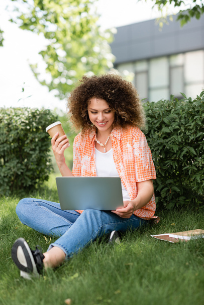 happy woman with curly hair holding paper cup and using laptop while sitting on grass  - Foto, Bild