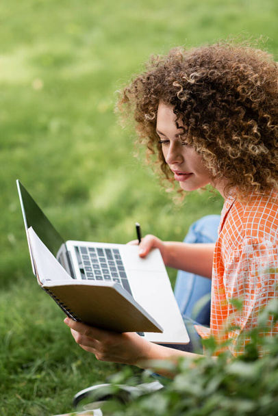 сфокусированная женщина с кудрявыми волосами читает заметки во время использования ноутбука в парке  - Фото, изображение