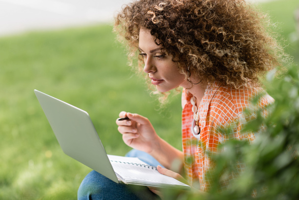 fokussierte Frau mit lockigem Haar benutzt Laptop, während sie Stift und Notizbuch im Park hält  - Foto, Bild