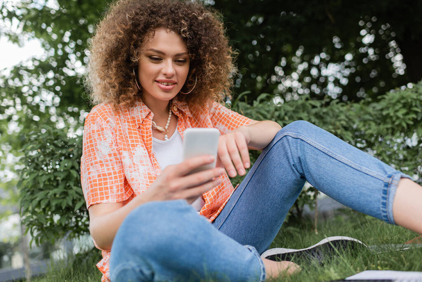 low angle view of positive freelancer woman with curly hair using smartphone in park  - Foto, imagen
