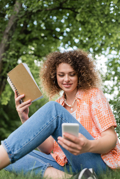 vista a basso angolo di giovane donna sorridente con riccio che tiene il taccuino durante l'utilizzo di smartphone nel parco verde  - Foto, immagini