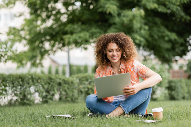 happy freelancer woman using laptop while sitting on green lawn near paper cup  - Foto, afbeelding