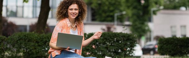 vrolijke freelancer vrouw met krullend haar houden laptop tijdens het werken op afstand in groen park, banner  - Foto, afbeelding