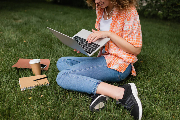 cropped view of happy woman using laptop while sitting on lawn near paper cup and notebook  - Foto, imagen