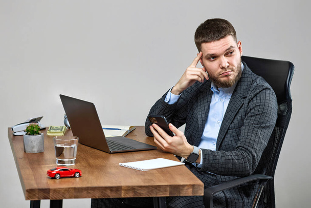 young bearded businessman sitting on chair at desk and holding phone, using laptop - 写真・画像