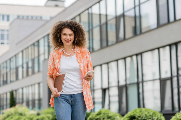 mujer alegre con pelo rizado sosteniendo teléfono inteligente y carpeta mientras camina fuera - Foto, imagen