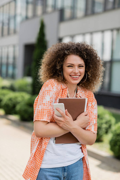 cheerful woman with curly hair holding smartphone and folder while smiling outside  - Foto, immagini
