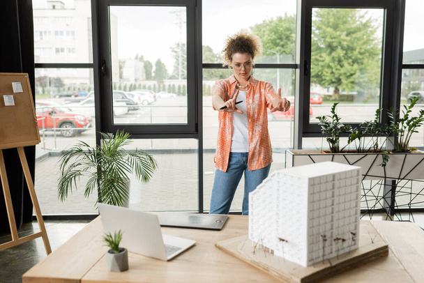 młody projektant robi gest ramki rękami patrząc na model domu mieszkalnego na biurku - Zdjęcie, obraz