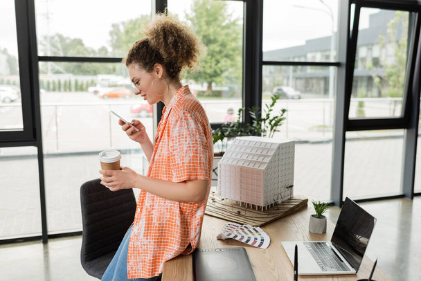 widok z boku szczęśliwy architektoniczny projektant w okularach trzymając kawę iść i za pomocą smartfona w biurze - Zdjęcie, obraz
