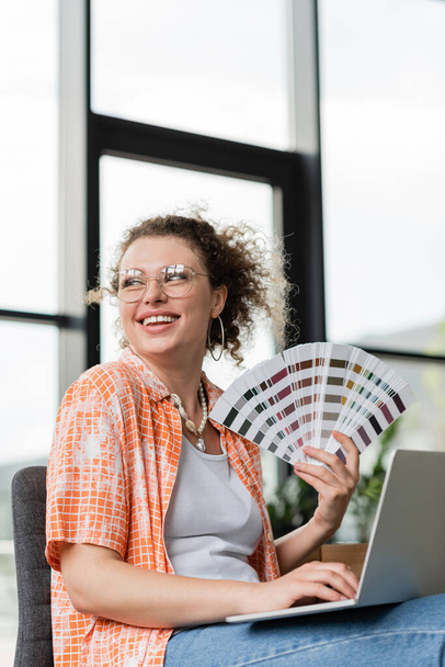 diseñador arquitectónico feliz en gafas que sostienen la paleta de colores cerca del ordenador portátil en la oficina - Foto, Imagen