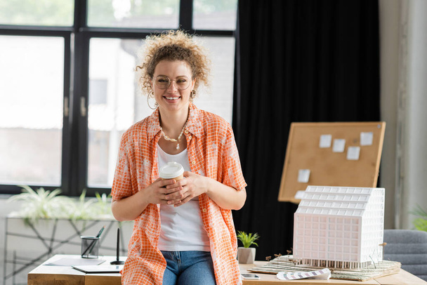 cheerful architectural designer holding paper cup while standing near house model in office  - Foto, imagen