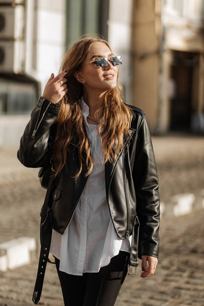 Modieus blond vrouw model met zwart leren jas en stijl zonnebril lopen de stad straat - Foto, afbeelding