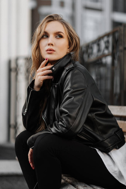 Retrato de cerca de una hermosa joven rubia sonriente en una chaqueta de cuero negro en el fondo de una calle de la ciudad - Foto, Imagen