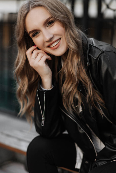 close-up portret van een mooie glimlachende jonge blonde vrouw in een zwart lederen jas op de een stad straat achtergrond - Foto, afbeelding
